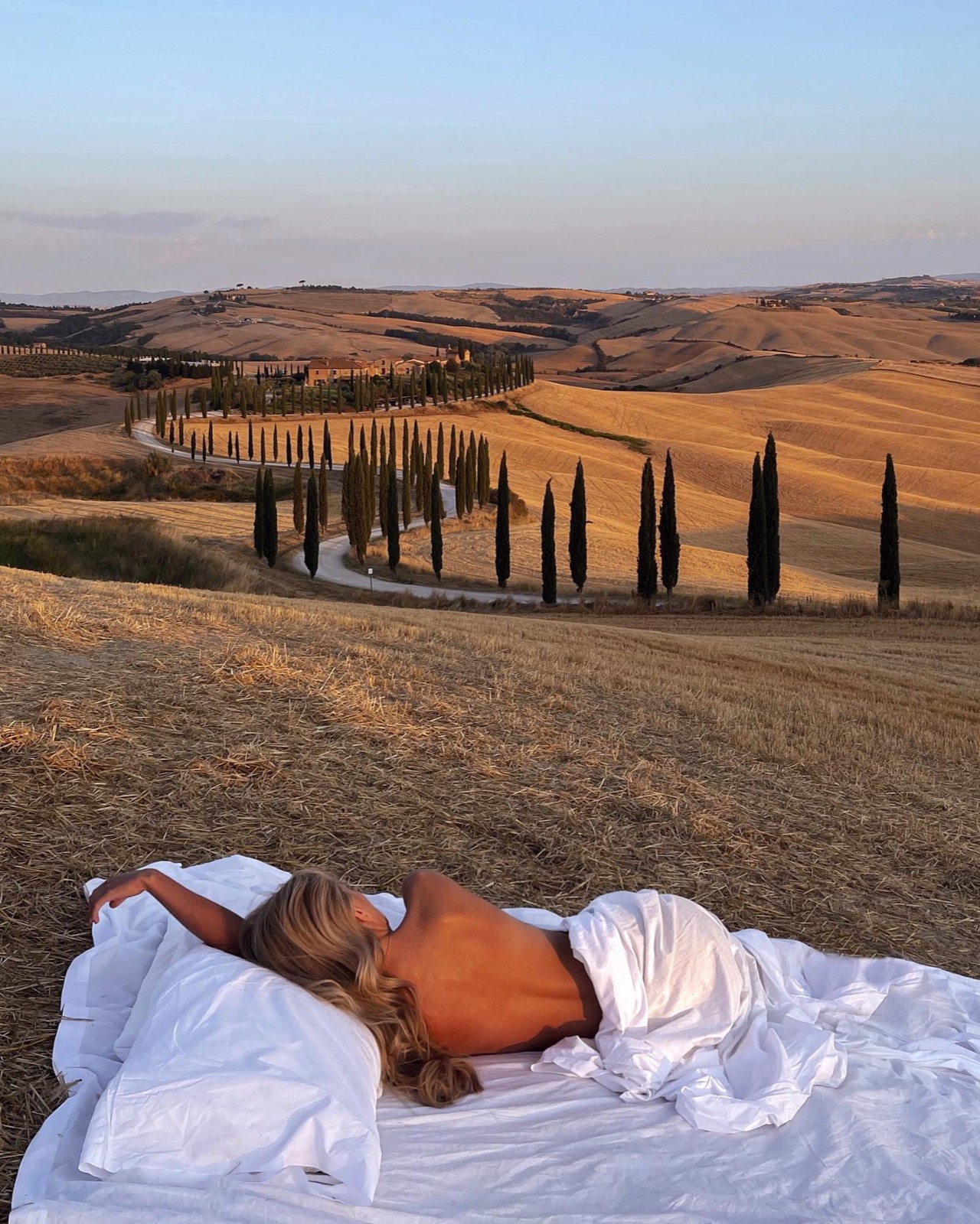 White linen bed set out above a cypress-lined road in Tuscany